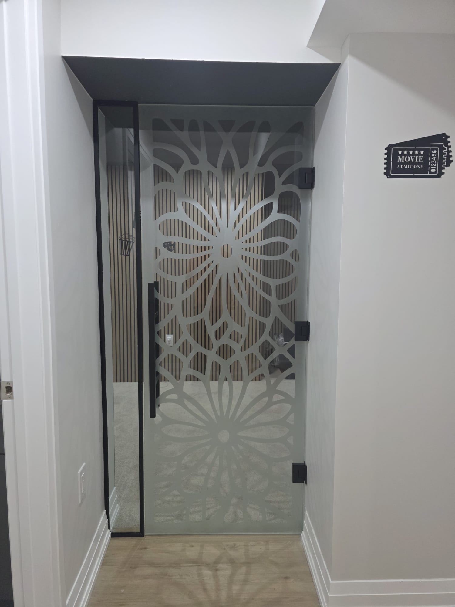 Custom acid-etched frameless glass door with intricate floral mandala pattern, matte black hinges, leading to home theatre room — Built By Glass, GTA