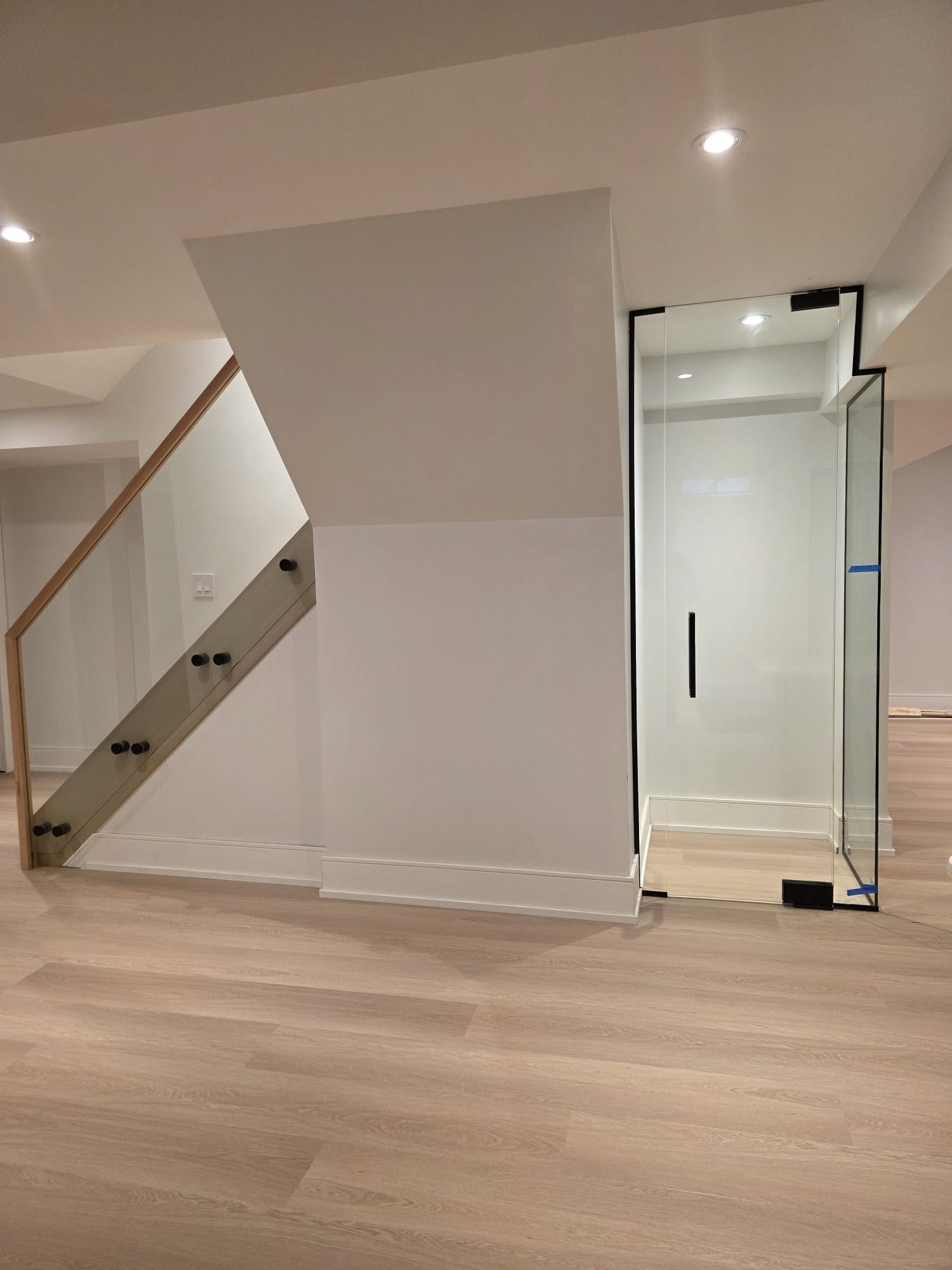 Frameless glass railing on basement staircase with light wood handrail and matte black standoff hardware, paired with frameless glass door enclosure — <a href=
